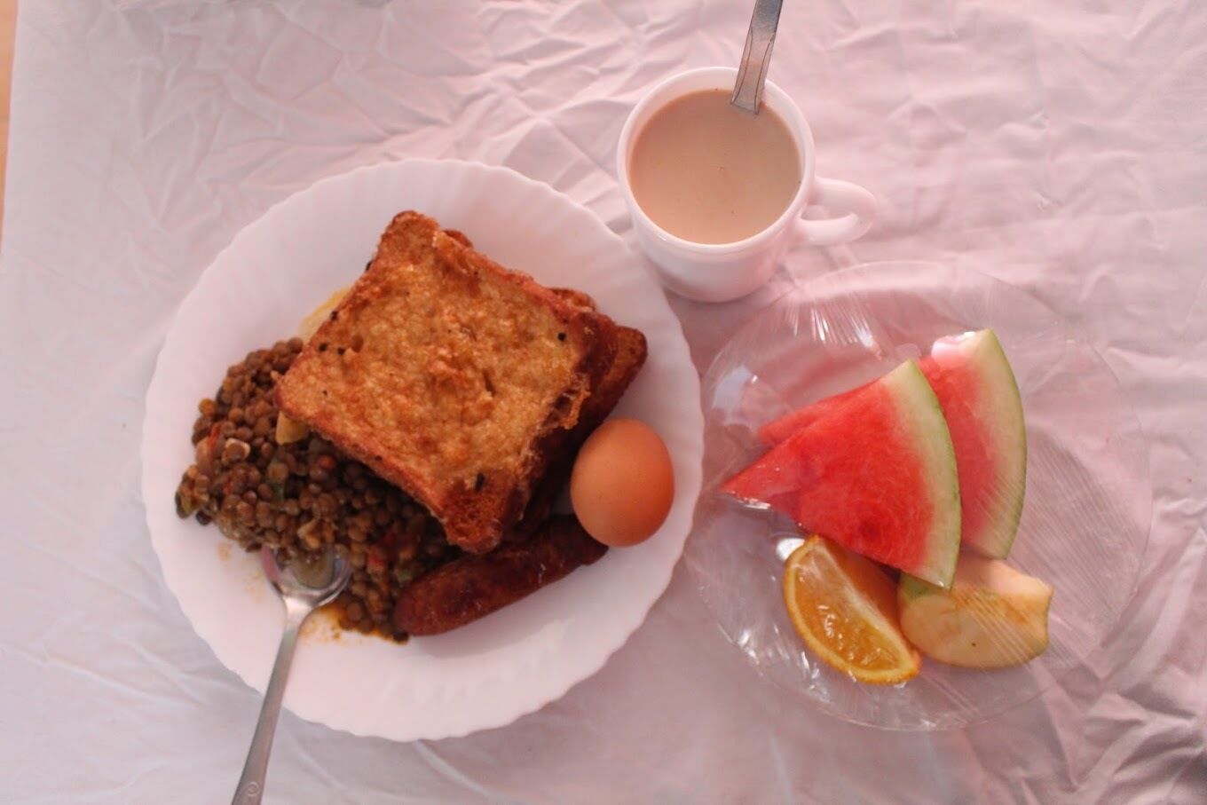 A breakfast plate with toast, lentils, an egg, and a cup of tea, along with a plastic plate containing watermelon and lemon slices.