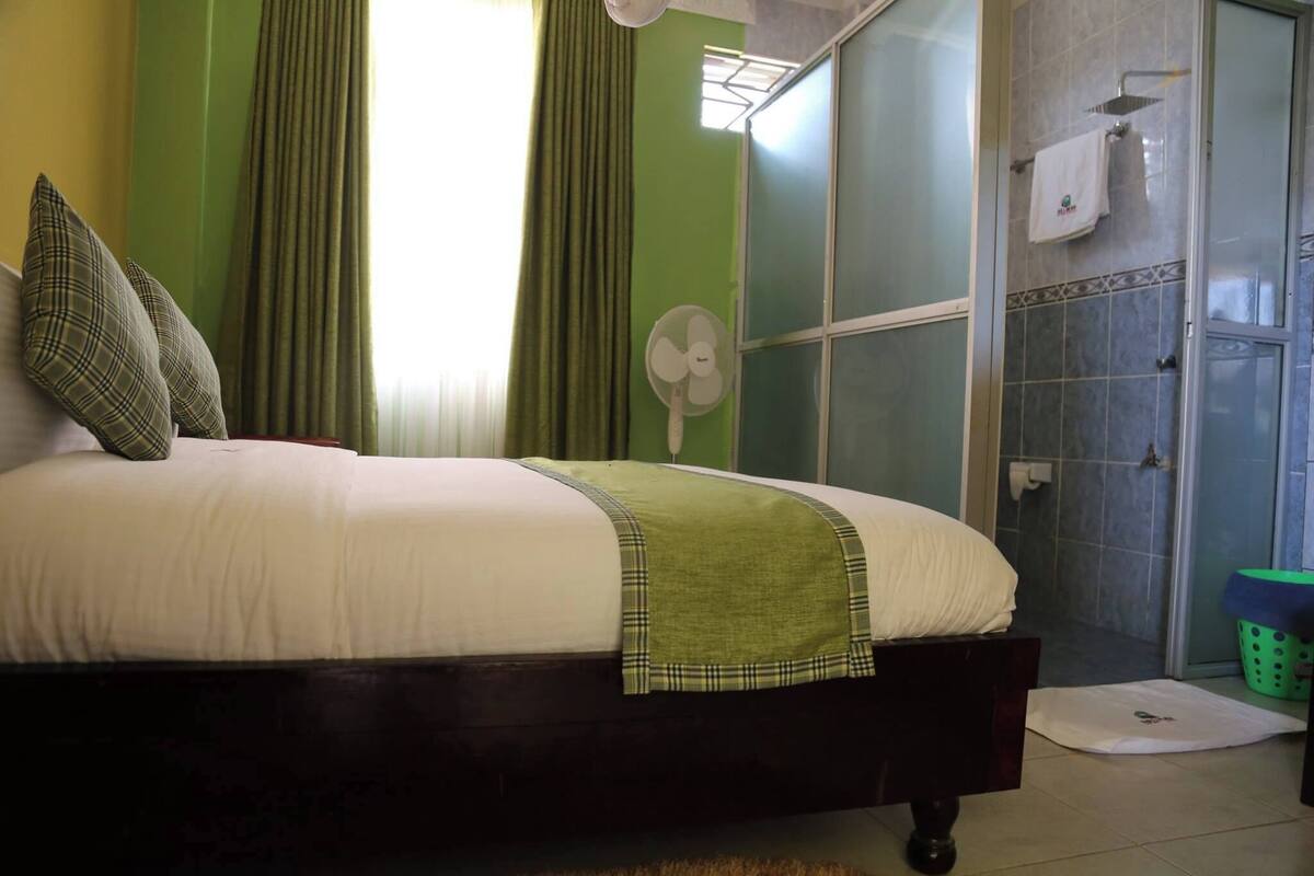 Un dormitorio con cama, cortinas verdes, un ventilador, zona de ducha y un cesto de ropa verde.