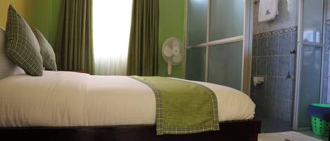A bedroom with a bed, green curtains, a fan, a shower area, and a green laundry basket.
