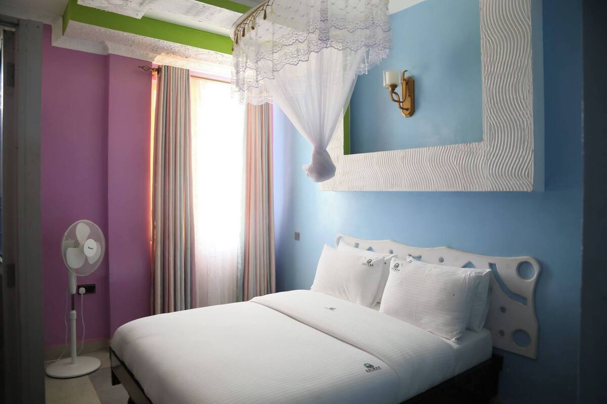 Un dormitorio con cama, ventilador de pie, ventana con cortinas y lámpara de pared.