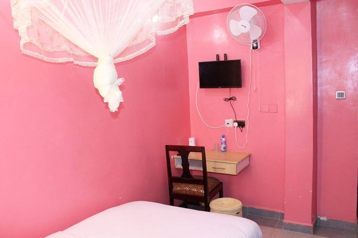 Una habitación rosa con una cama, un escritorio de madera, una silla, una mesita de noche, un ventilador y un televisor colgado en la pared.