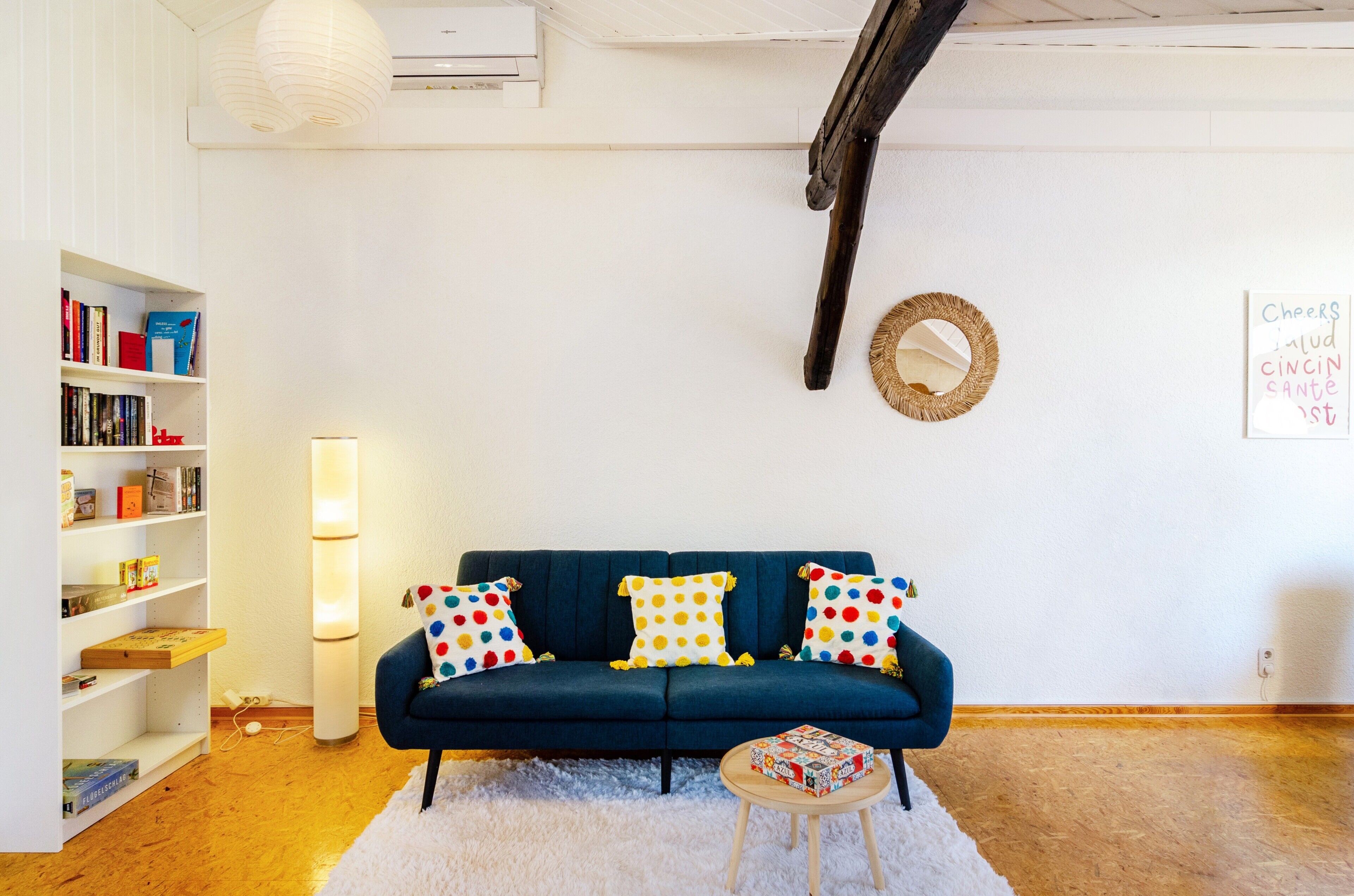Een gezellige woonkamer met een donkerblauwe bank, kleurrijke stippenkussens, een witte boekenkast, een ronde spiegel en een wit vloerkleed.