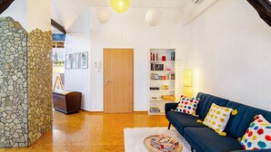 A cozy living room with a stone wall, a wooden door, a bookshelf, a blue sofa with polka dot cushions, and a small round table with a board game. - Apartment 'Stadt-Loft' with Private Terrace, Wi-Fi, and Air Conditioning (Nordhausen)
