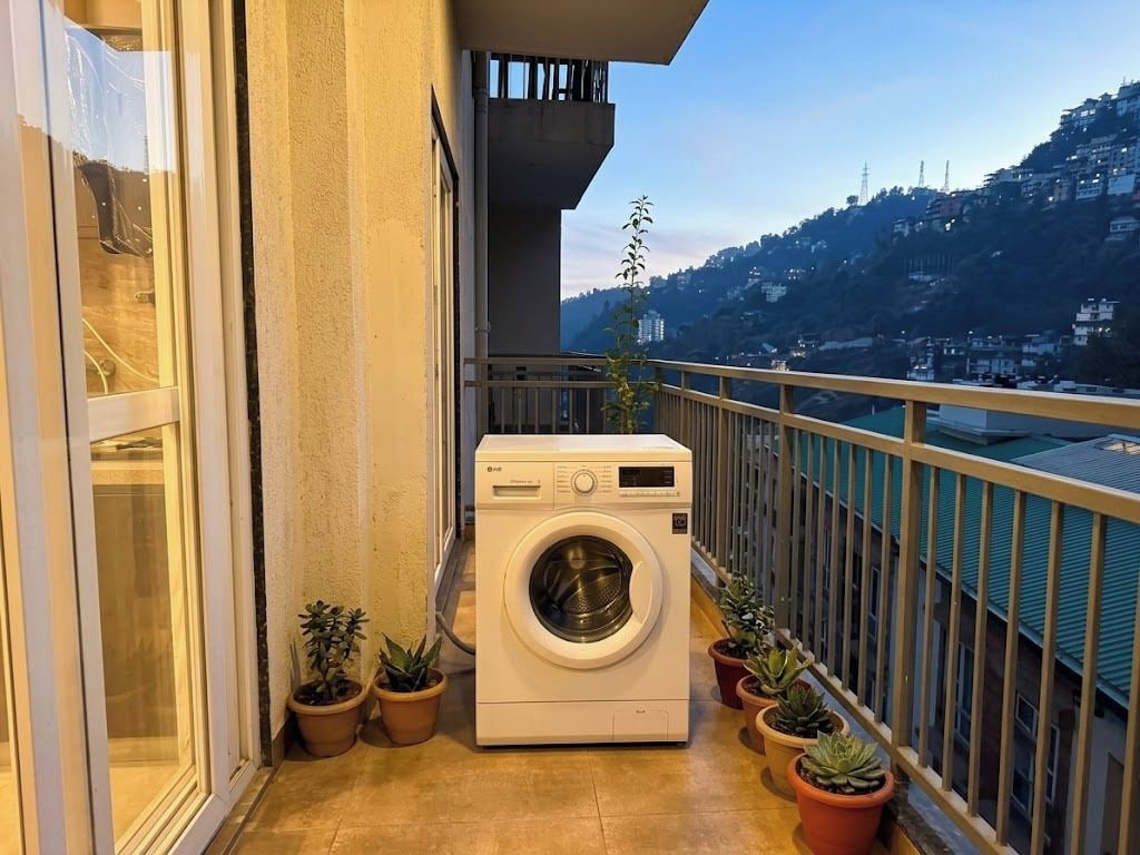 A balcony with a washing machine, potted plants, and a view of a hillside.
