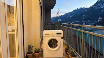 A balcony with a washing machine, potted plants, and a view of a hillside.