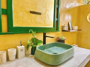 Casa de banho com lavatório verde, espelho, prateleira com artigos de higiene e uma planta em vaso.