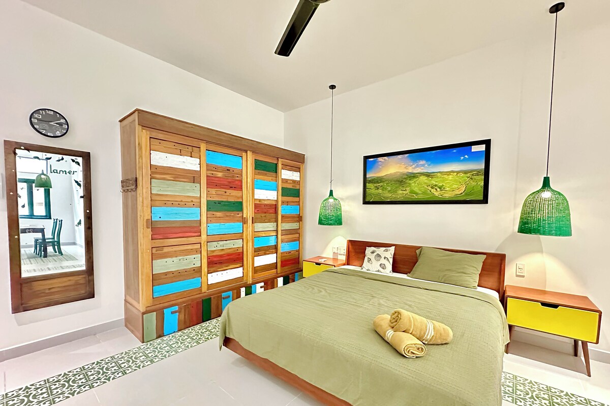 A bedroom with a bed, a wooden wardrobe, a painting, and two green pendant lights.