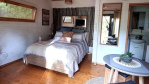 Une chambre confortable avec un plafond en bois, un lit recouvert d’une couette grise et blanche, un ventilateur de plafond, une petite table avec une plante et une fenêtre avec des rideaux.