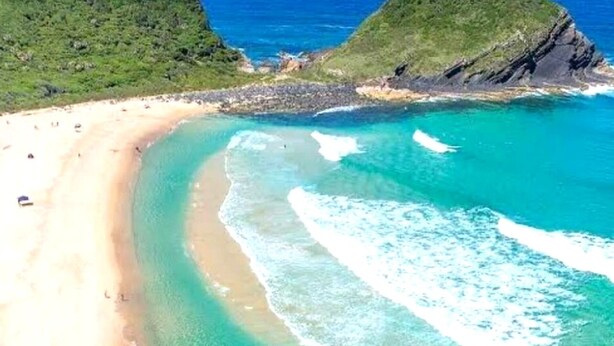 Une plage côtière aux eaux turquoise et limpides, bordée de sable fin, avec des collines verdoyantes en arrière-plan.