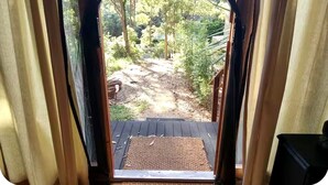 Vue depuis une pièce dont la porte en bois donne sur un espace extérieur comprenant une terrasse en bois, un paillasson et un jardin avec des arbres et un sentier.