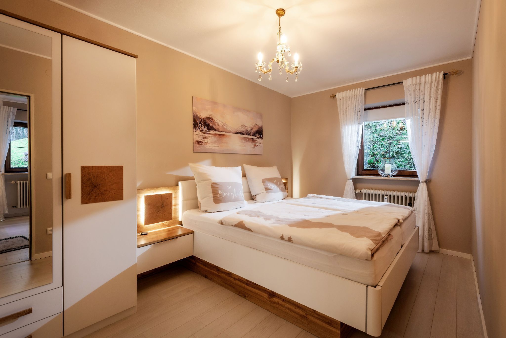 Une chambre à coucher avec un lit, des tables de chevet, un lustre, une fenêtre avec des rideaux et un tableau au mur.
