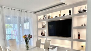 Un comedor moderno con una mesa larga, sillas blancas y un televisor montado en una pared blanca con estantes debajo.