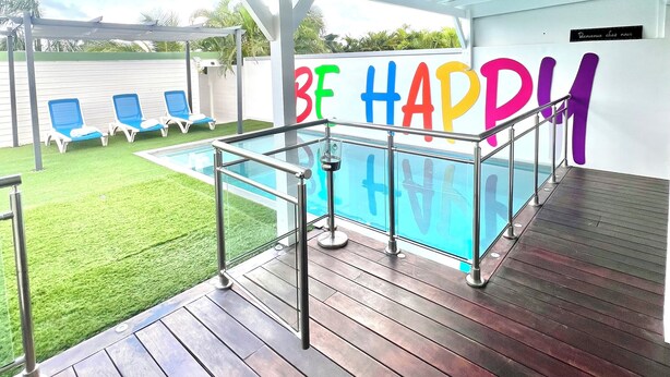 Un área de piscina techada con cubierta de madera, baranda de vidrio y un cartel que dice 'BE HAPPY'.