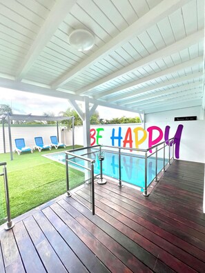 A covered pool area with a wooden deck, a glass railing, and a sign that reads "BE HAPPY". - Jacuzzi, Pool & Sea View in Orient Bay (Saint-Martin)