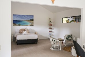 A bedroom with a bed, a chair, a shelf with books, and a painting of a beach. - Studio on Percheron: Minimal. Mindful. Artful (Margaret River)