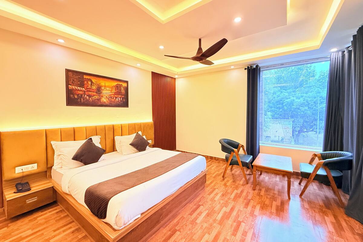 A modern hotel room with a large bed, a ceiling fan, a painting on the wall, and a window with a view of trees.