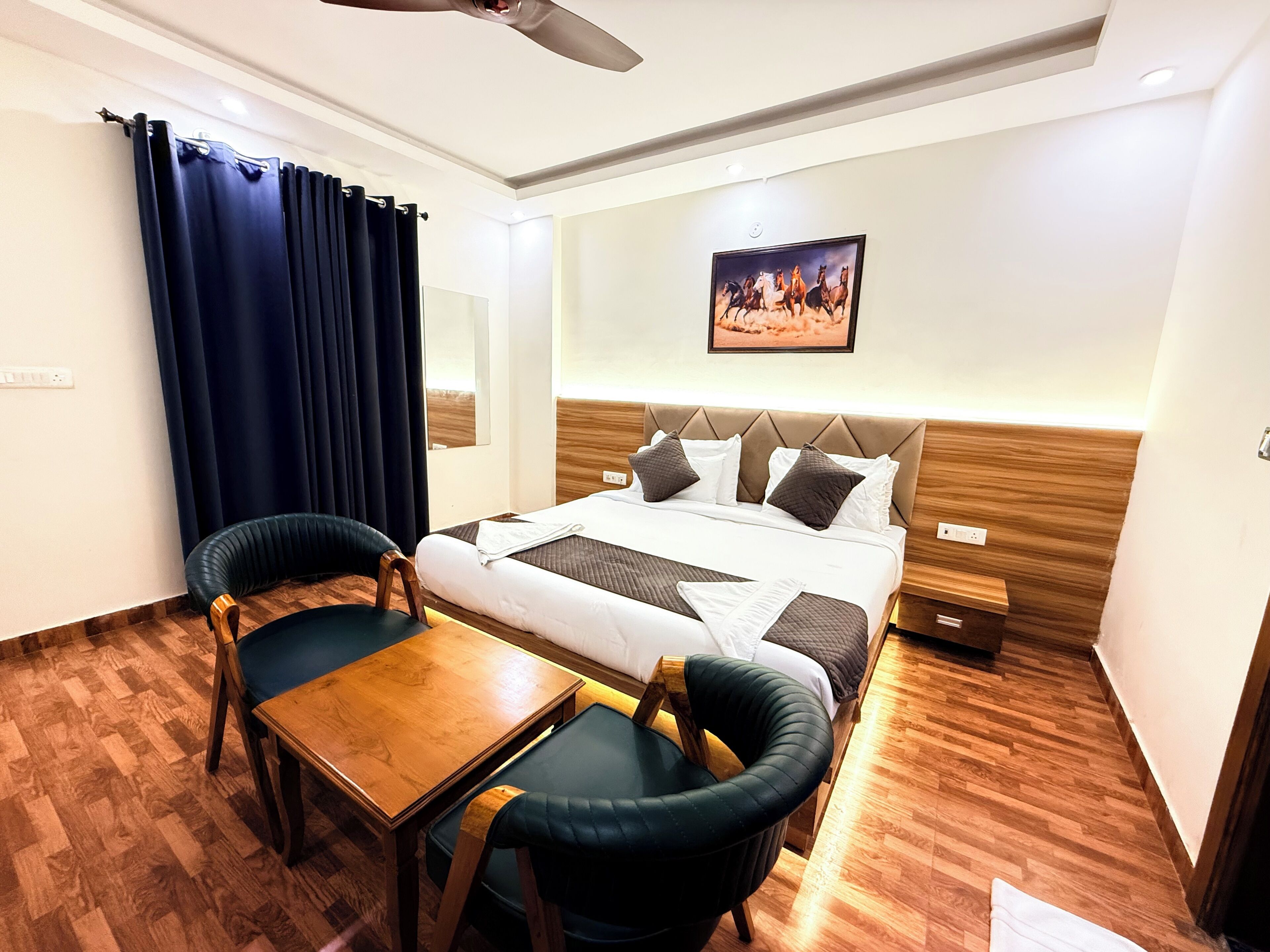 A hotel room with a bed, two chairs, a table, a ceiling fan, a painting, and a wooden floor.