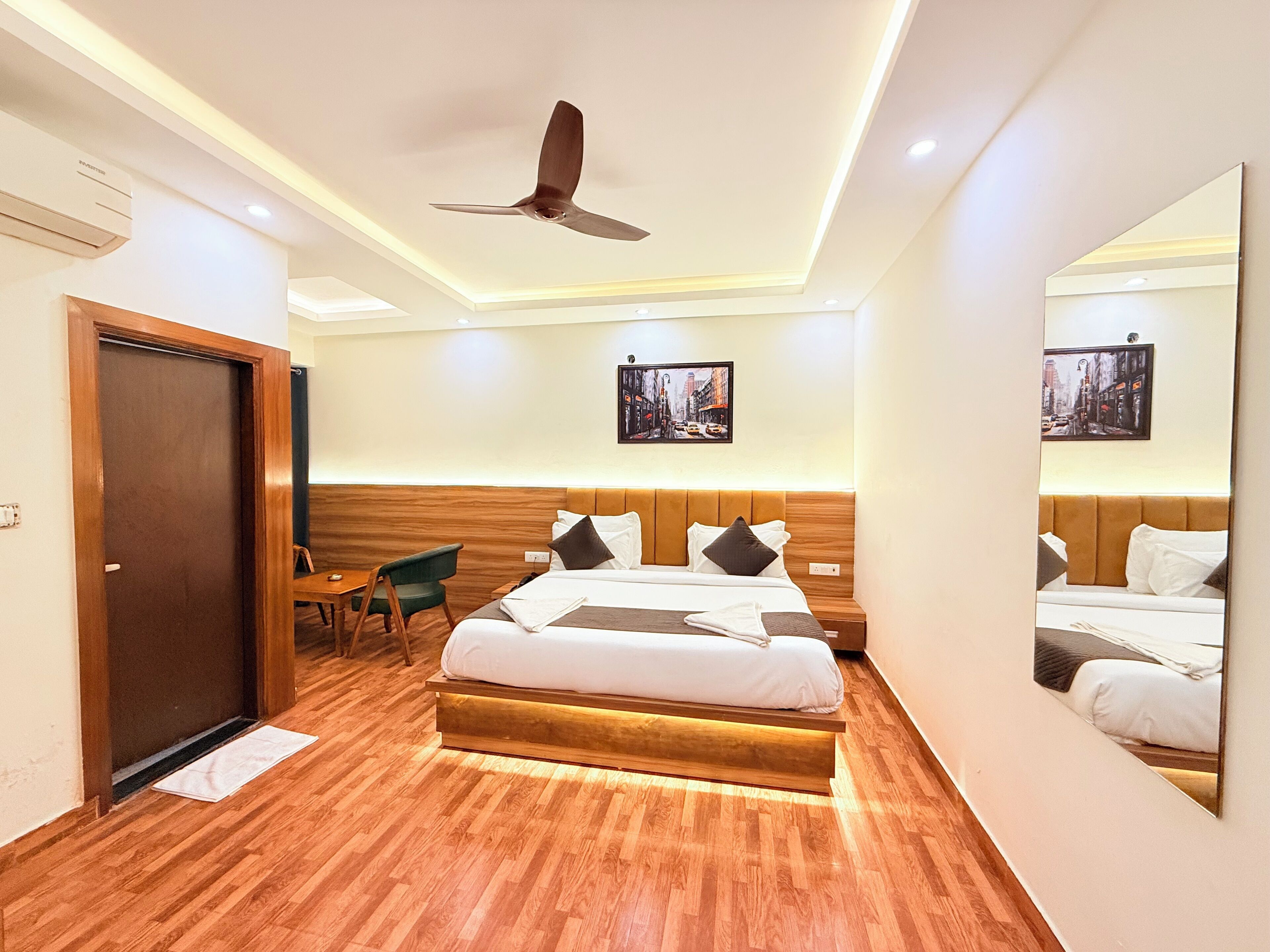 A modern hotel room with a wooden floor, a bed with white bedding, a desk with a chair, a ceiling fan, and two framed pictures on the wall.