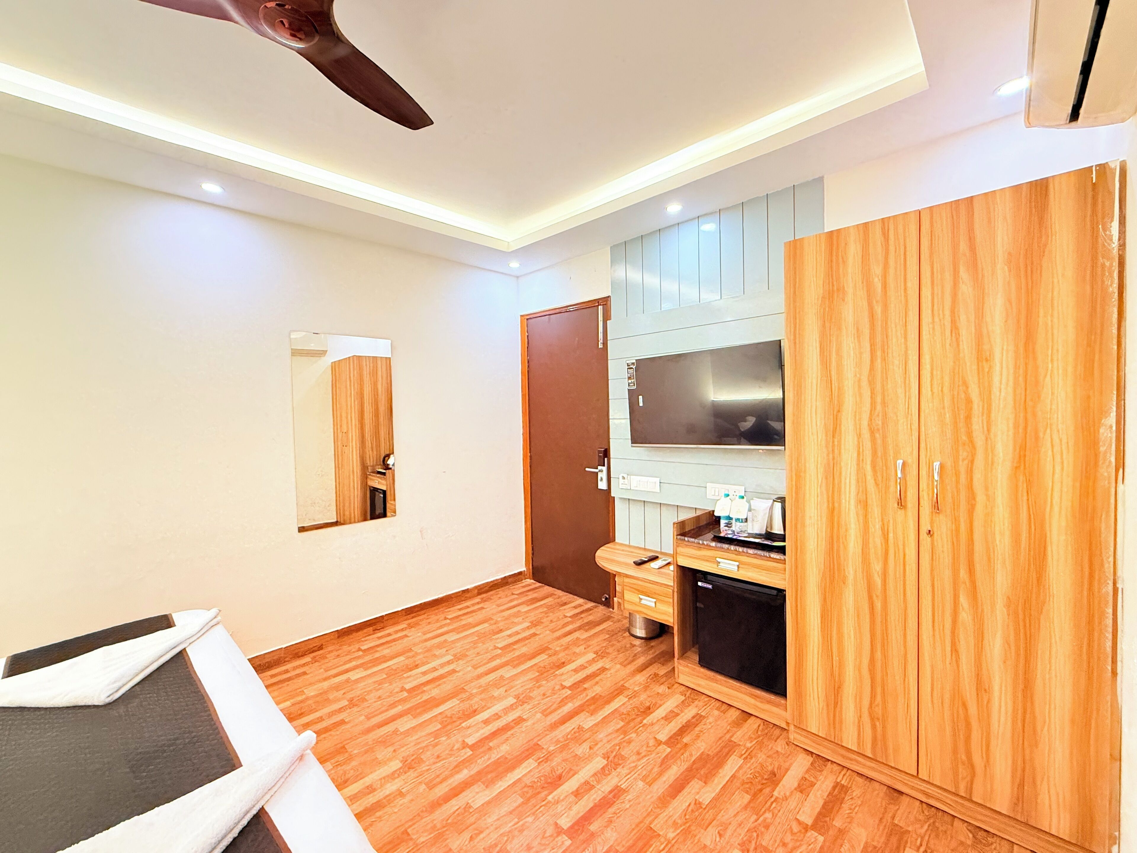 A modern hotel room with a wooden floor, a ceiling fan, a large wardrobe, a small kitchenette, and a mirror.