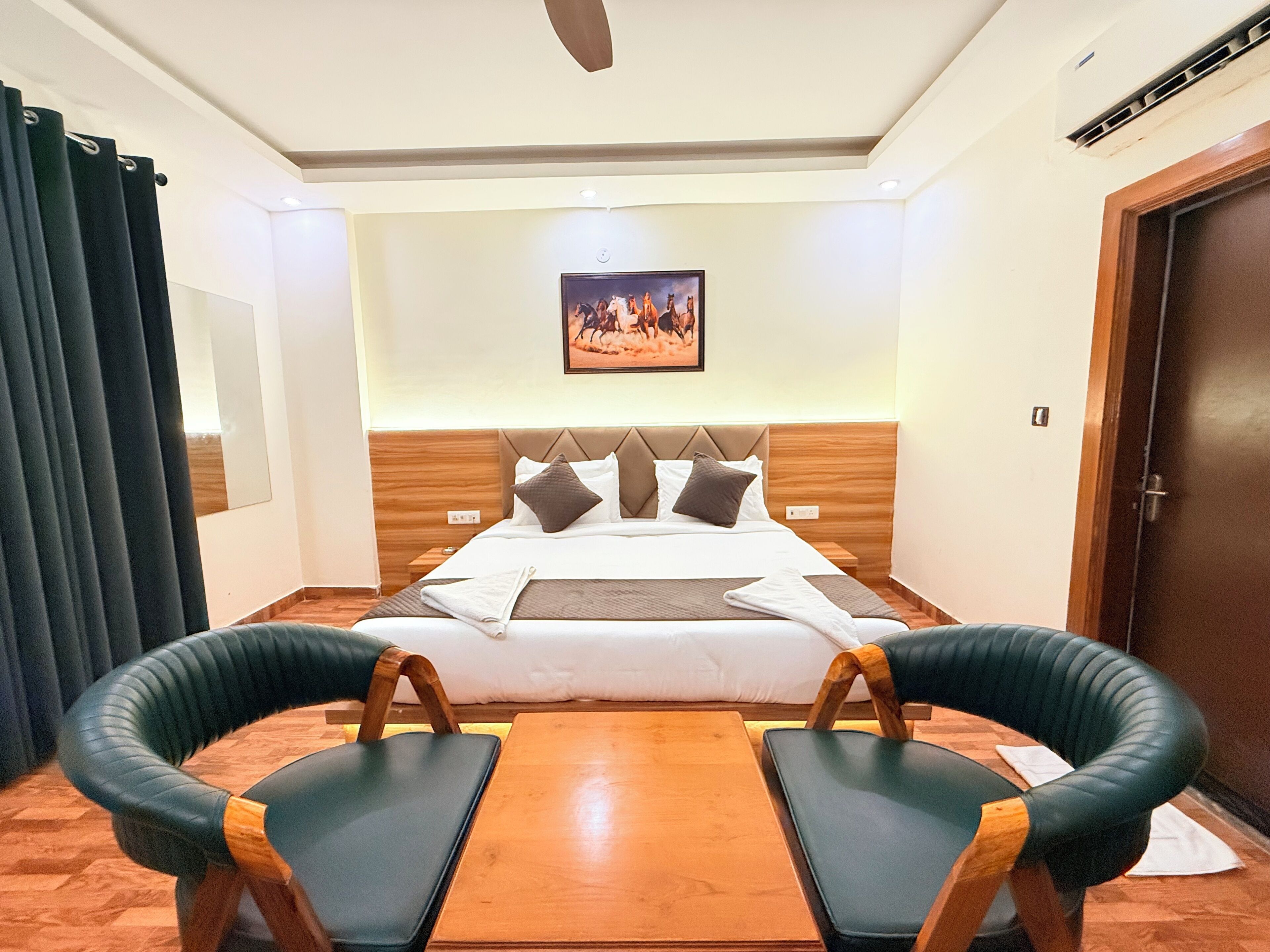 A hotel room with a bed, two armchairs, a wooden table, a ceiling fan, and a painting on the wall.