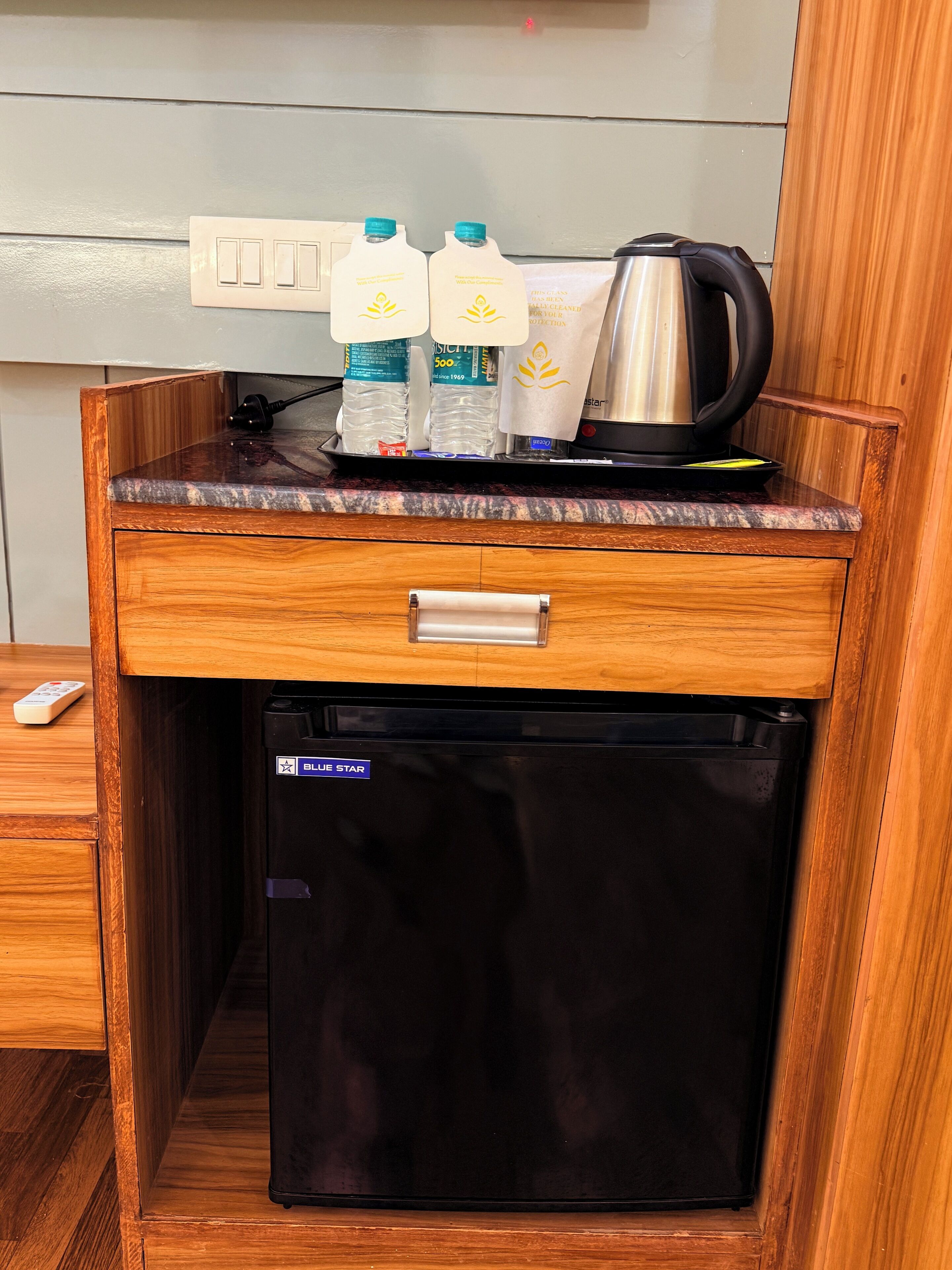 A compact kitchenette with a wooden countertop, a small refrigerator, a coffee maker, and two water bottles.