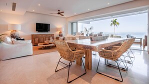 Un salon spacieux avec une grande table à manger en bois, des chaises en osier, un canapé blanc et un téléviseur fixé au mur.