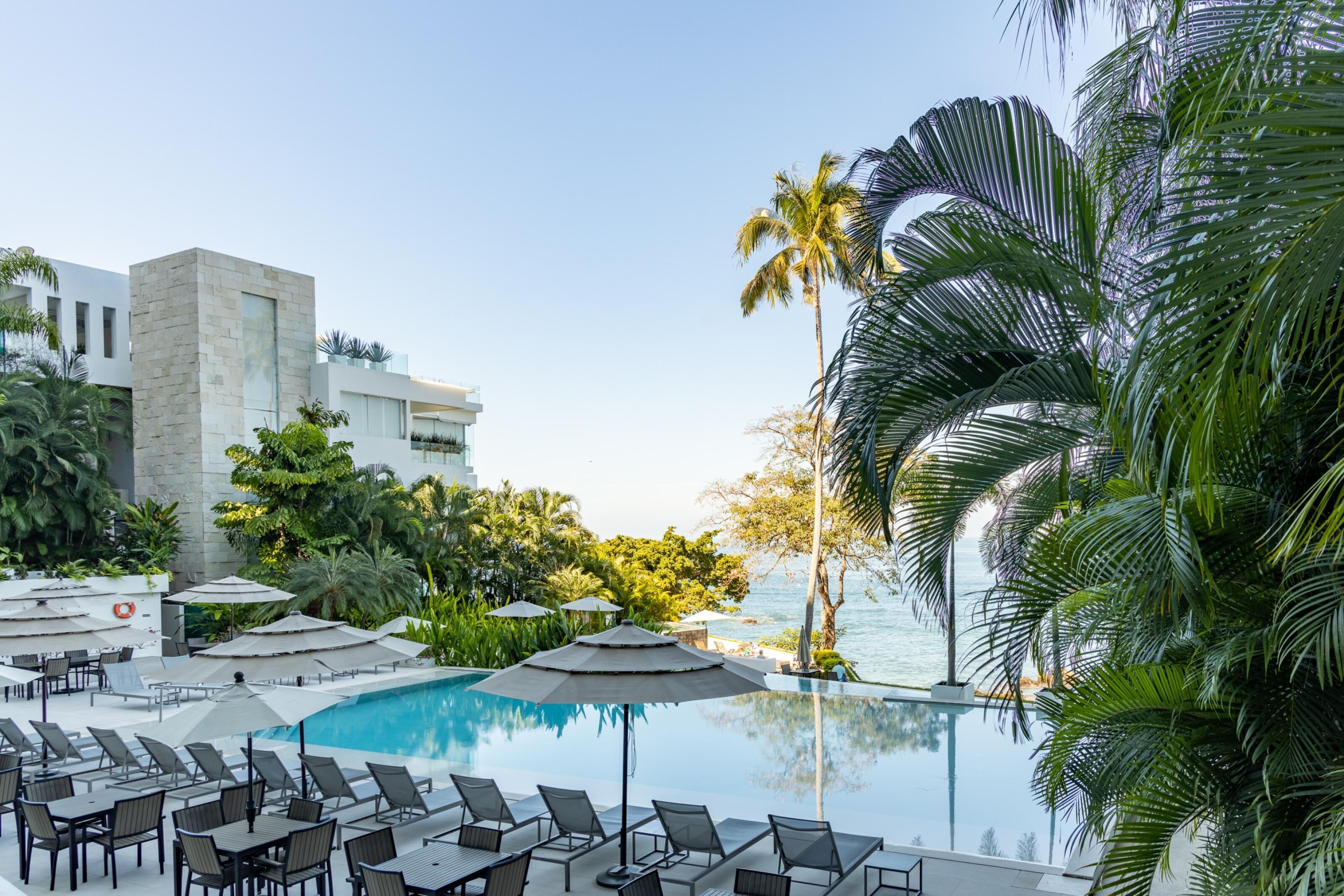 Un espace aménagé au bord de la piscine, avec des chaises longues et des parasols, une vue dégagée sur l’océan et un bâtiment moderne en arrière-plan.