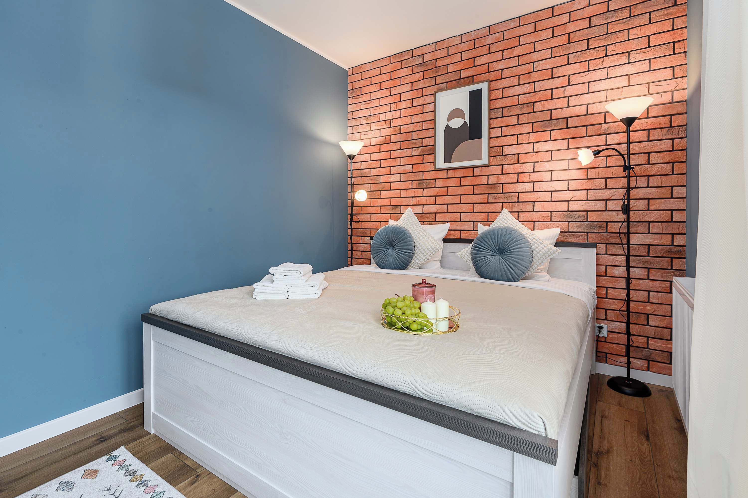 A bedroom with a brick-patterned wall, a bed with white bedding, a bedside table with a lamp, and a framed picture above the bed.