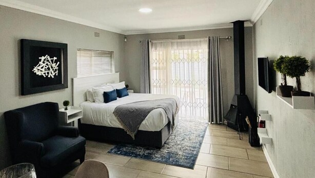 A modern bedroom with a bed, bedside table, chair, and a large window with curtains. - Rolem Guest House (Randburg)