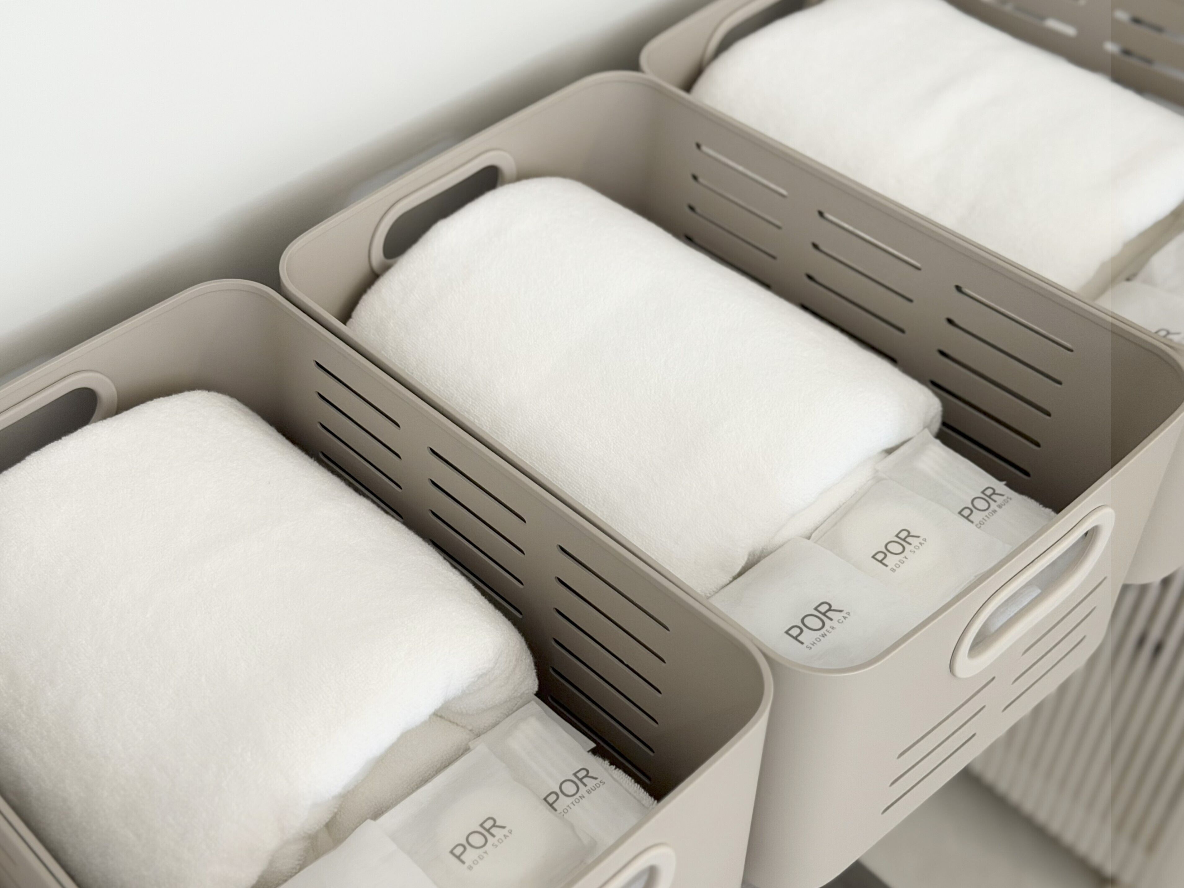 A set of gray storage bins with white towels and labeled soap packets.
