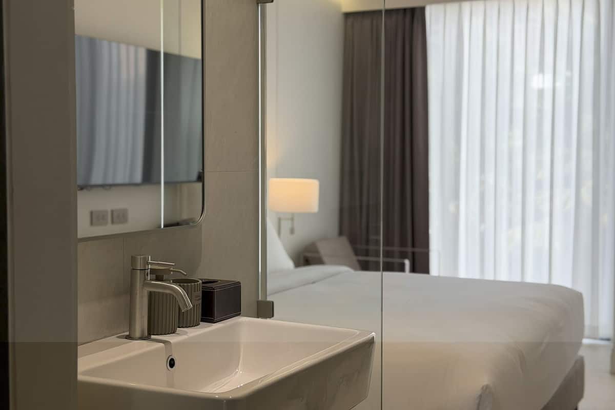 A hotel room with a large bed, a bathroom with a sink and mirror, and a window with curtains.