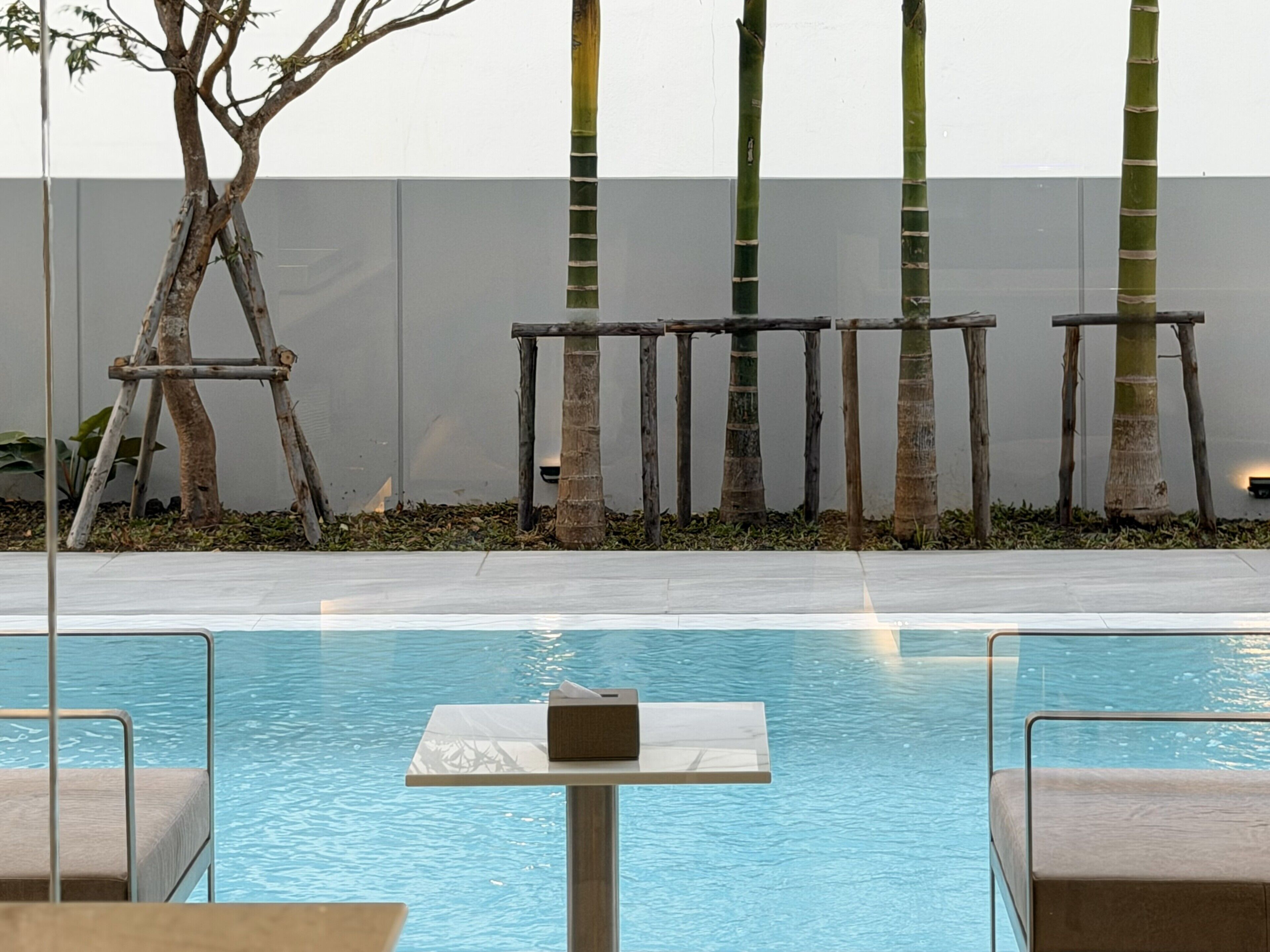 A pool area with a table and chairs, surrounded by bamboo plants and a white wall.
