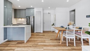 A modern kitchen with white cabinetry, a stainless steel refrigerator, and a wooden dining table with white chairs. - Olympic Park Oasis | 2BR + Pool, Sauna & Parking (Sydney Olympic Park)