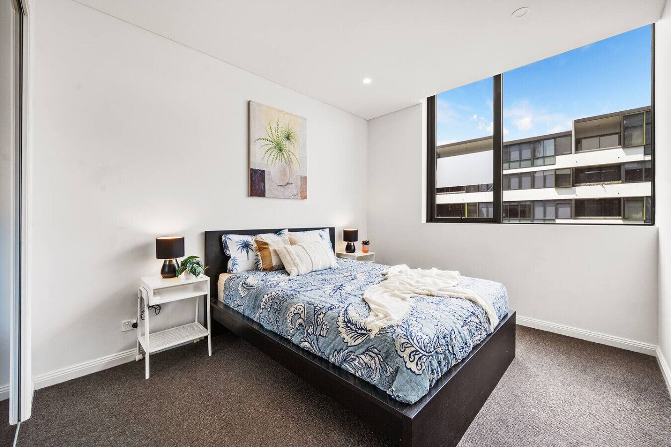 A modern bedroom with a large bed, white nightstands, a single artwork, and a view of a building through a large window.