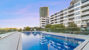 A swimming pool with a glass barrier, surrounded by modern buildings and greenery. - Olympic Park Oasis | 2BR + Pool, Sauna & Parking (Sydney Olympic Park)