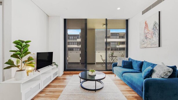 A modern living room with a blue sofa, a flat-screen TV, a coffee table, and a potted plant. - Olympic Park Oasis | 2BR + Pool, Sauna & Parking (Sydney Olympic Park)