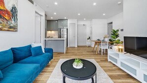 A modern apartment with a blue sofa, a dining area, a kitchen, and a TV. - Olympic Park Oasis | 2BR + Pool, Sauna & Parking (Sydney Olympic Park)