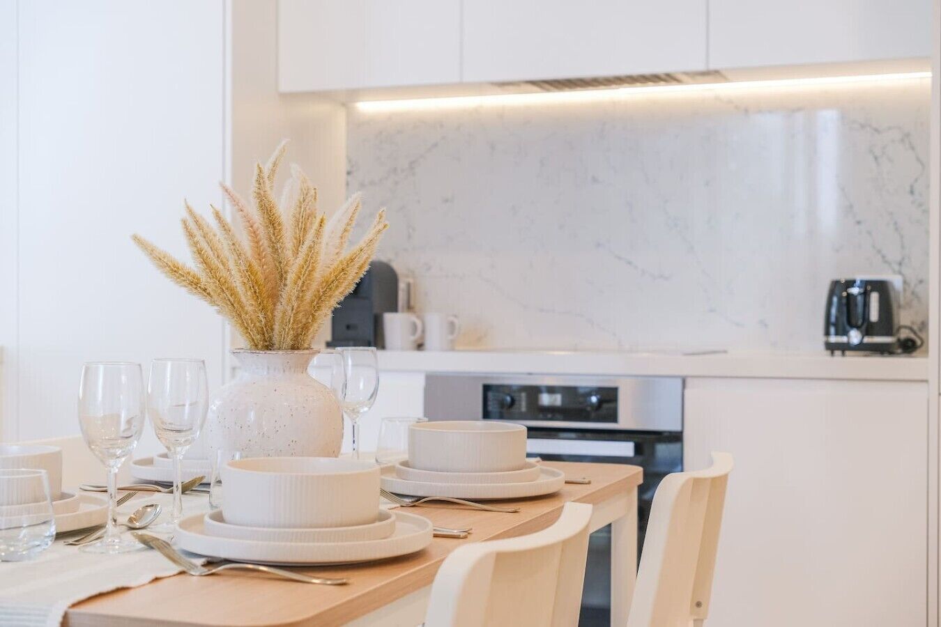A modern kitchen with white cabinetry, a marble countertop, and a dining table set with white plates, glasses, and cutlery.