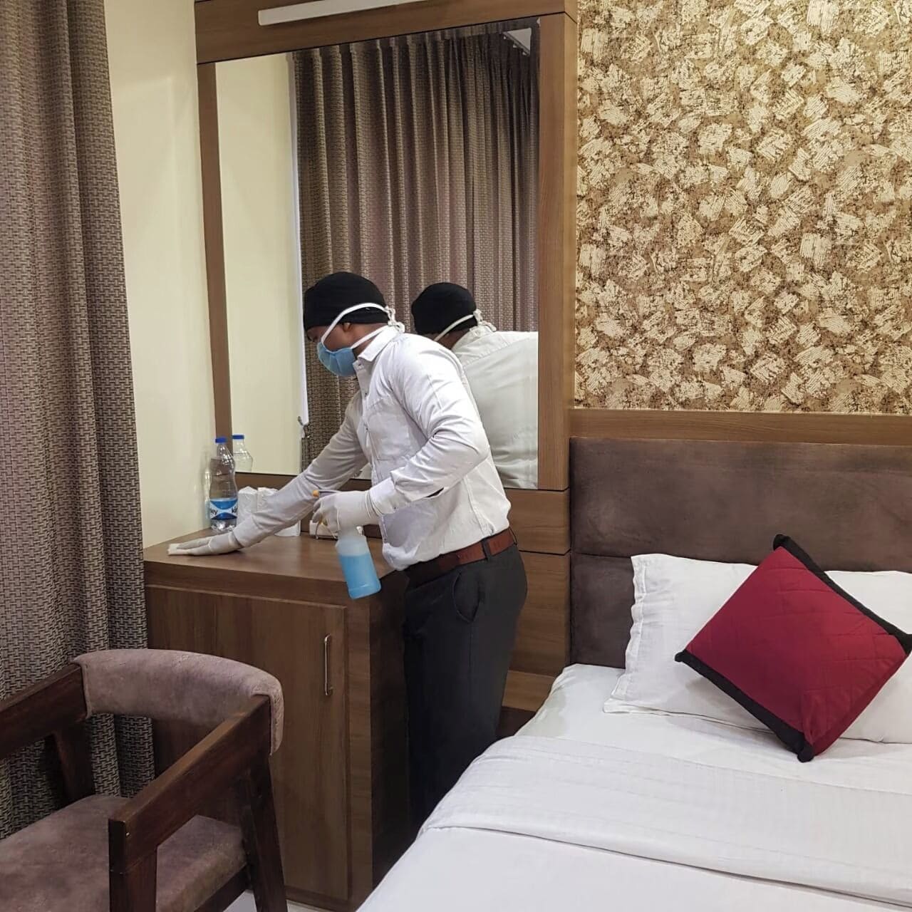 A hotel room with a wooden desk, a mirror, a bed with white bedding and a red pillow, and a chair.