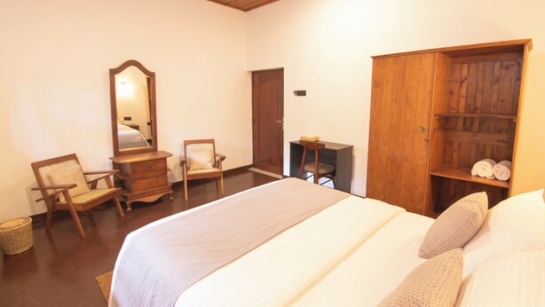 A bedroom with a large bed, wooden wardrobe, two chairs, a dresser, and a mirror. - Sand Cliff Villa (Galle)
