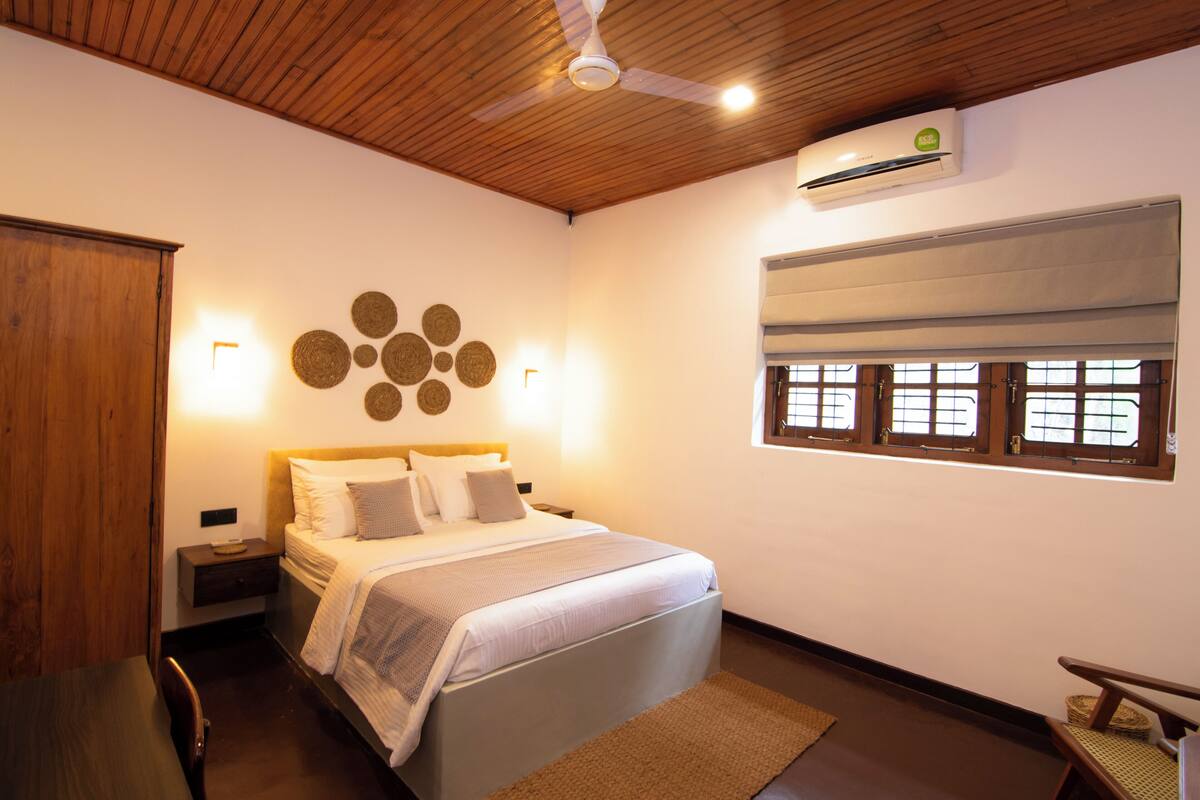 A bedroom with a bed, bedside tables, a wooden ceiling, an air conditioning unit, a window with blinds, and a wall decoration.