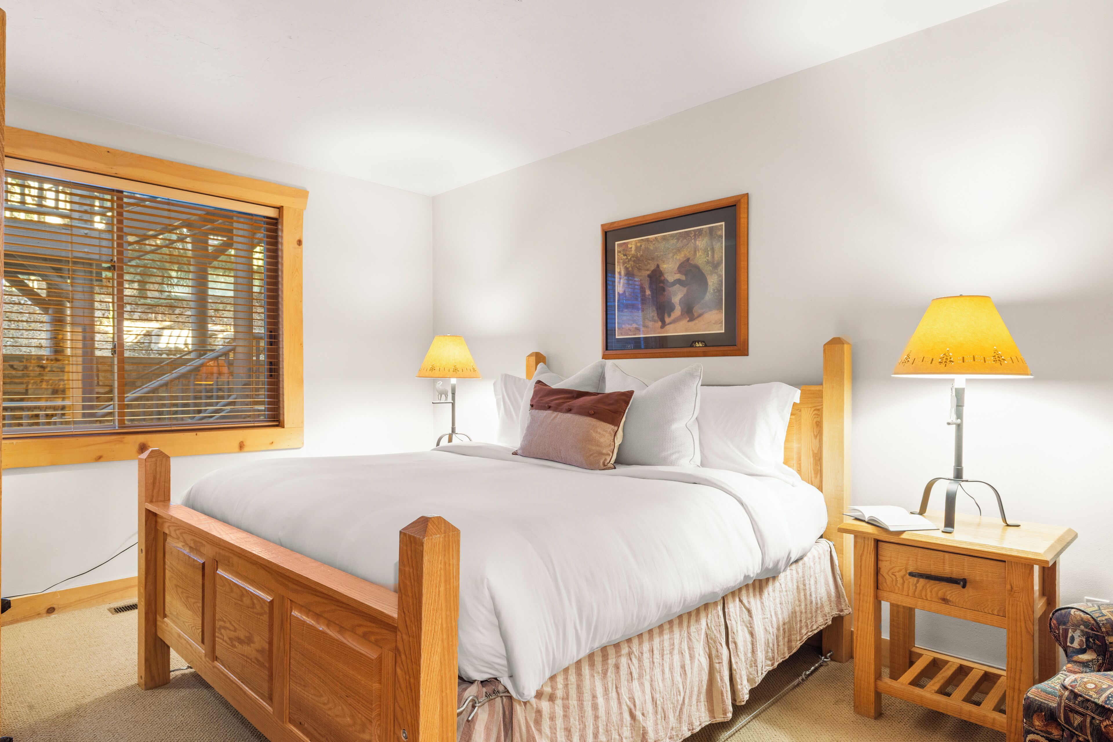 A neatly arranged bedroom with a wooden bed, two bedside tables with lamps, a framed picture on the wall, and a window with blinds.