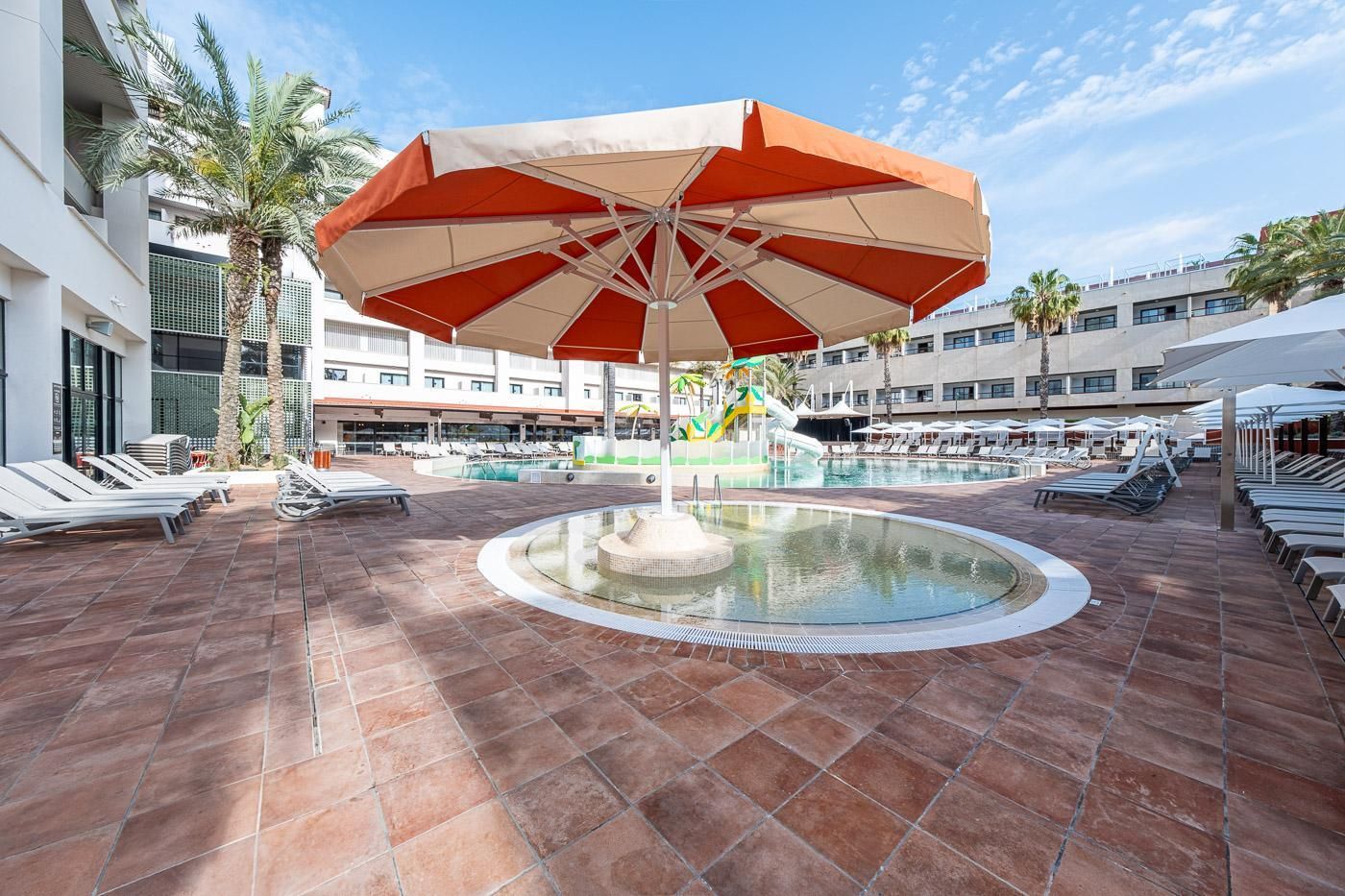 Un área de piscina con sillones de descanso, una fuente central y una sombrilla grande.