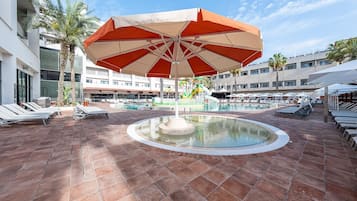 Un área de piscina con sillones de descanso, una fuente central y una sombrilla grande.