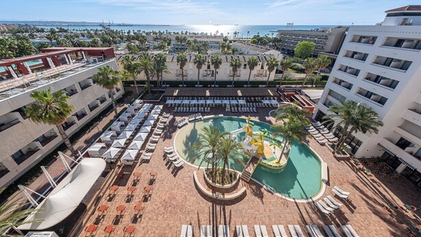 Un espace piscine aménagé sur le toit, avec de nombreux chaises longues et parasols, entouré de palmiers et d’immeubles.