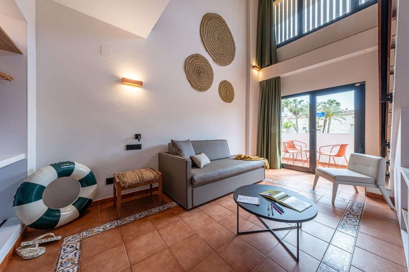 A modern living room with a grey sofa, a small wooden stool, a round coffee table, and a large decorative rug. There are two wall-mounted lights and three woven wall decorations.