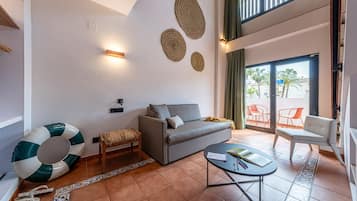 A modern living room with a grey sofa, a small wooden stool, a round coffee table, and a large decorative rug. There are two wall-mounted lights and three woven wall decorations.