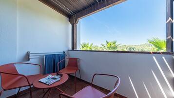 A balcony with two red metal chairs, a small round table with a magazine, and a view of greenery and hills.