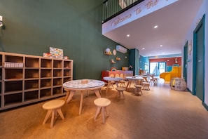Un espace de jeux pour enfants avec des tables et des tabourets ronds, une bibliothèque et un panneau d’accueil.