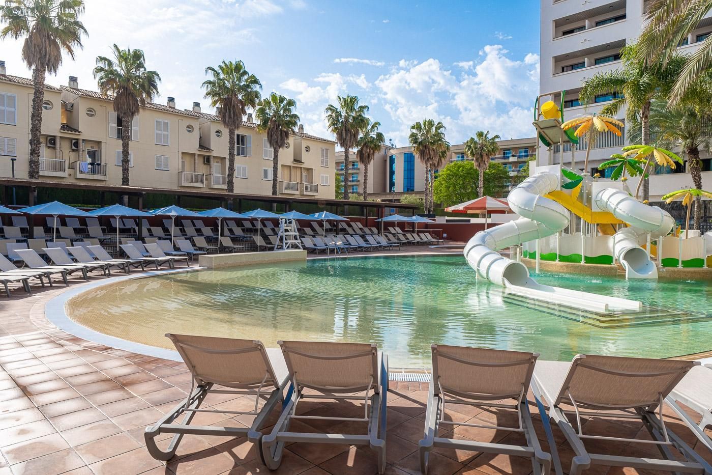 A pool area with lounge chairs, a water slide, and palm trees.